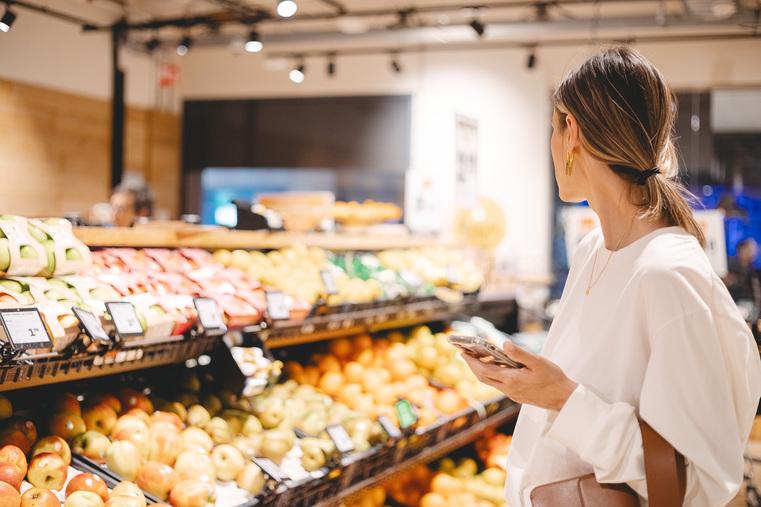 Frau mit Smartphone im Supermarkt an Obstauslage. Visualisiert Kältetechnik und Klimasysteme für den Einzelhandel.