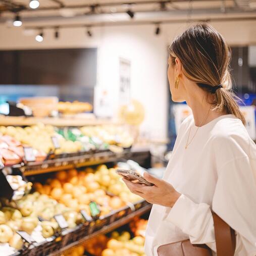 Frau mit Smartphone im Supermarkt an Obstauslage. Visualisiert Kältetechnik und Klimasysteme für den Einzelhandel.