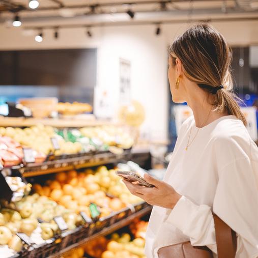 Frau mit Smartphone im Supermarkt an Obstauslage. Visualisiert Kältetechnik und Klimasysteme für den Einzelhandel.