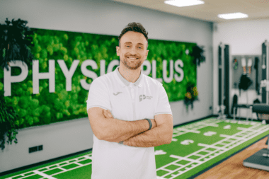 Trainer l&auml;chelt in Physiotherapie-Praxis mit Kunstrasen. Stellt Klimasysteme und L&uuml;ftungstechnik f&uuml;r Sportst&auml;tten dar.